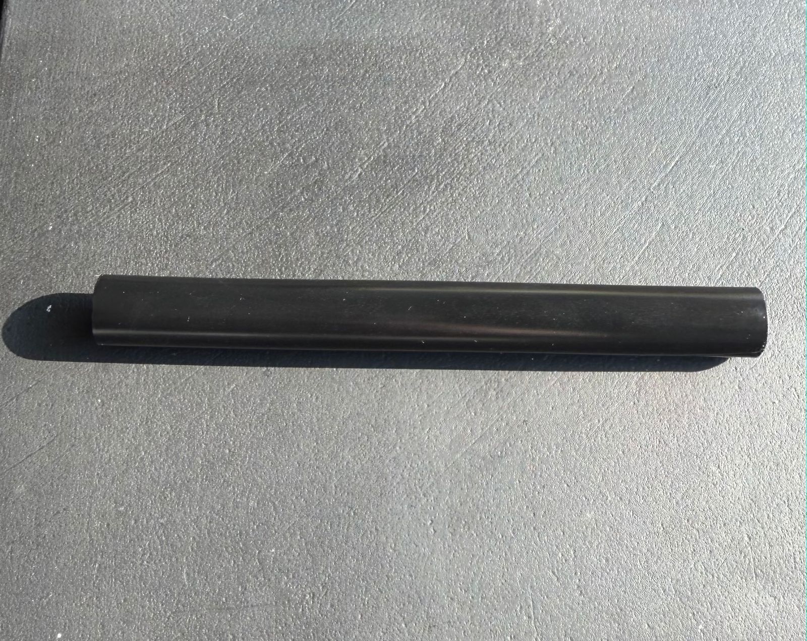 The Buddy System Safety Grip on its own: a straight black tube lying on a dark, textured surface in sunlight.